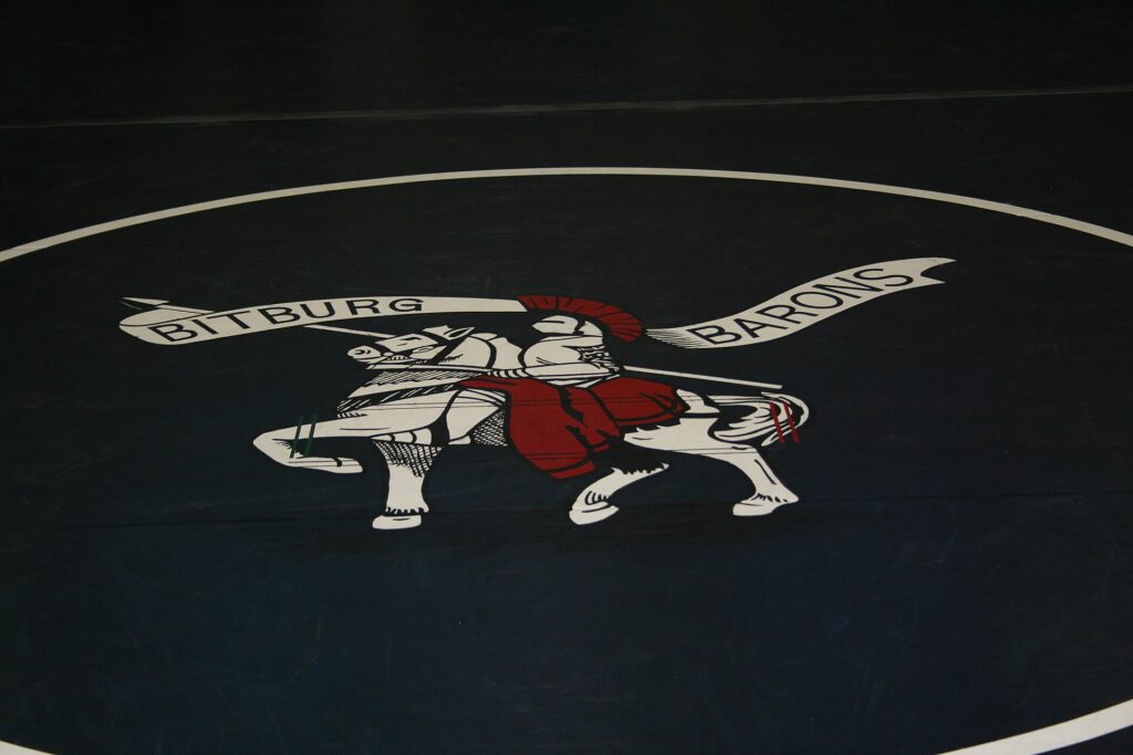 Two wrestlers competing on a circular mat inside a hall with officials seated alongside