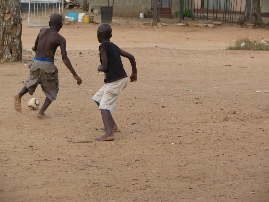 Children playing football on a paved street between parked cars and low walls