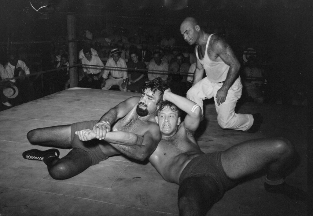 A referee crouching beside two wrestlers to watch the hold at close range