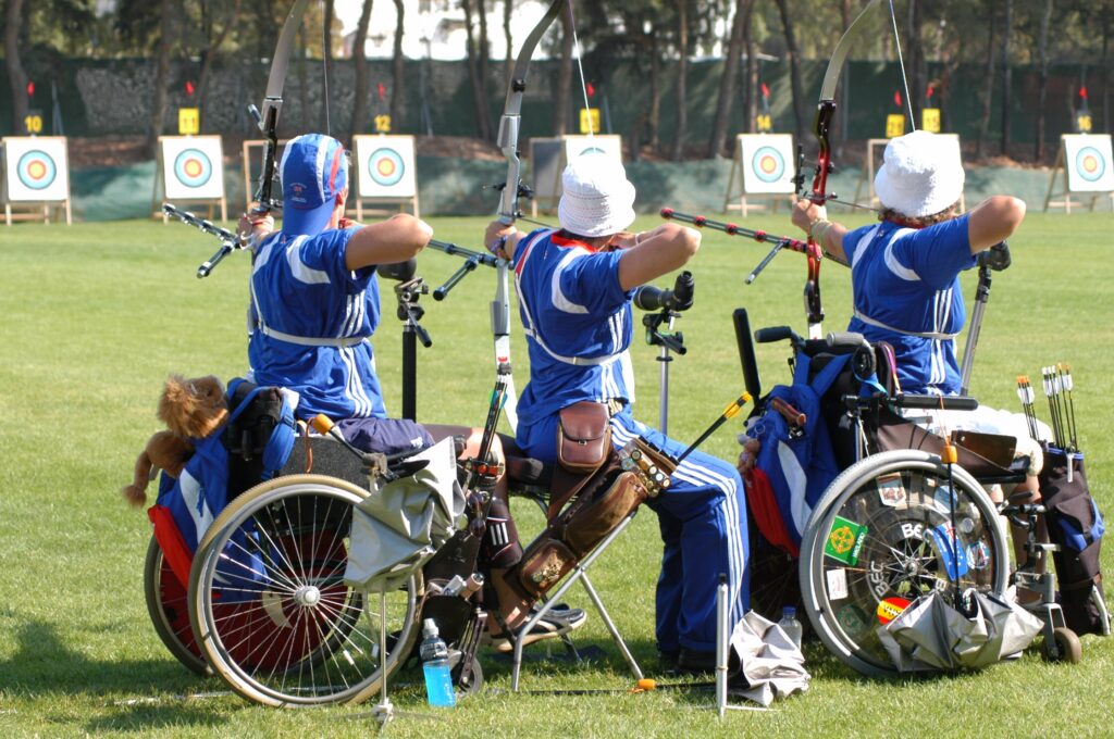 A seated archer aiming a recurve bow from a wheelchair on a grass shooting range