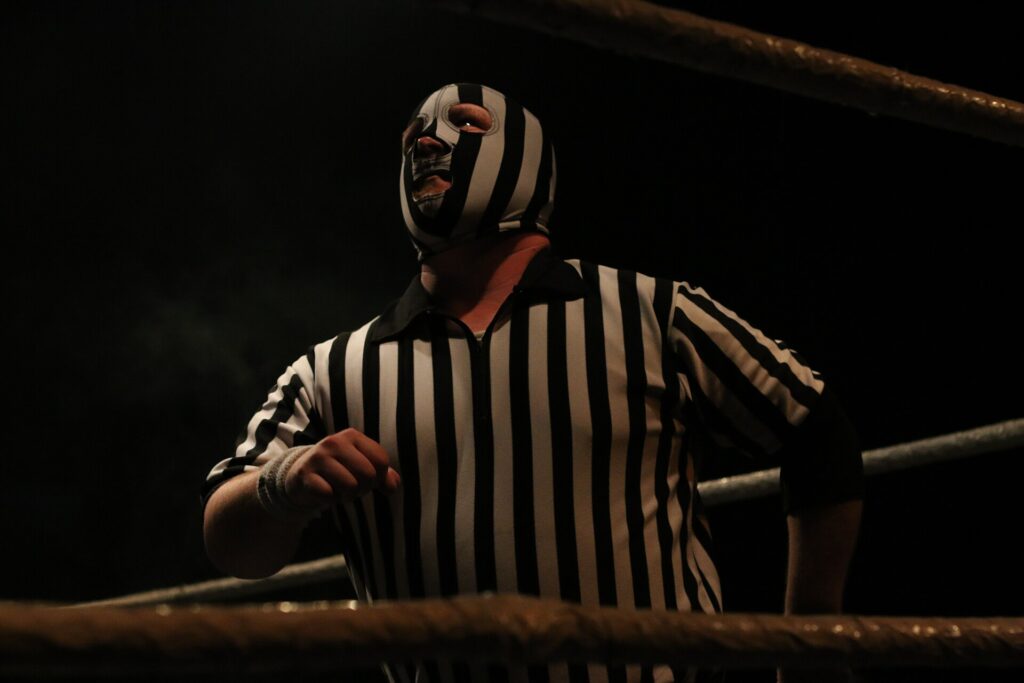 A referee in uniform raising an arm to signal a decision beside two wrestlers on the mat