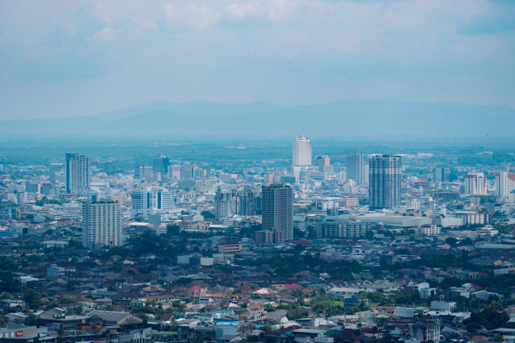 A city spread below snow-topped mountains seen from a road on the slope above it